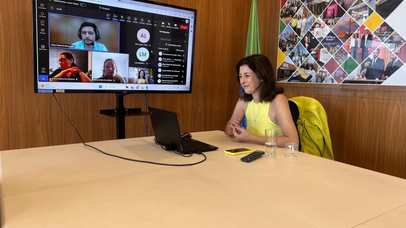Foto da secretária sentada à mesa em frente ao computador. Ao seu lado, uma tv exibe os demais participantes da reunião. 