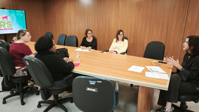 Vista ampla da mesma sala de reuniões, com seis mulheres sentadas em torno da mesa. A mulher à esquerda segue falando, enquanto as outras acompanham. A parede ao fundo é de madeira e o lado direito da sala tem janelas grandes com vista para o exterior. Há sofás e um ar-condicionado no canto.