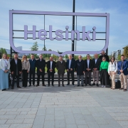 A foto mostra um grupo de 16 pessoas posando em frente a um grande letreiro que diz "Helsinki". Eles estão em uma área aberta com piso de pedra, cercada por grama e árvores, com prédios modernos ao fundo. A maioria está vestida de forma formal, com ternos e blazers, enquanto alguns usam roupas casuais, como jeans e uma jaqueta verde. O céu está claro, sugerindo um dia ensolarado.