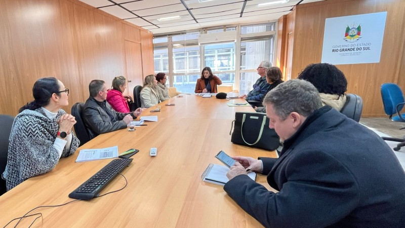 Vista de frente da mesma reunião, com a mesa ocupada por participantes dos dois lados. A mulher que lidera a fala está ao fundo, de frente para os demais. À direita da imagem, há uma bolsa sobre a mesa e um homem de casaco preto escreve em um caderno. Ao fundo, há janelas grandes com luz natural e o brasão do Governo do Estado do Rio Grande do Sul fixado na parede de madeira clara.