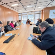 Vista de frente da mesma reunião, com a mesa ocupada por participantes dos dois lados. A mulher que lidera a fala está ao fundo, de frente para os demais. À direita da imagem, há uma bolsa sobre a mesa e um homem de casaco preto escreve em um caderno. Ao fundo, há janelas grandes com luz natural e o brasão do Governo do Estado do Rio Grande do Sul fixado na parede de madeira clara.