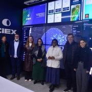 Grupo de nove pessoas posa em uma sala com grandes telas digitais ao fundo. As telas exibem mapas, gr&aacute;ficos e o nome &ldquo;CIEX&rdquo;, em um ambiente tecnol&oacute;gico de monitoramento. O grupo est&aacute; alinhado lado a lado, voltado para a c&acirc;mera, em frente a um painel com dados e imagens relacionadas a eventos clim&aacute;ticos.