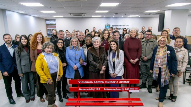 Grupo numeroso de pessoas posa em uma sala institucional do Governo do Estado do Rio Grande do Sul, atr&aacute;s de um banco vermelho com a mensagem: &ldquo;Viol&ecirc;ncia contra mulher &eacute; crime! Denuncie! 190 &ndash; Informa&ccedil;&otilde;es: 180&rdquo;.
Os participantes est&atilde;o organizados em v&aacute;rias fileiras para uma foto oficial, em ambiente iluminado, com o bras&atilde;o do Estado vis&iacute;vel na parede ao fundo.