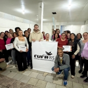 Descri&ccedil;&atilde;o da imagem: Grupo de aproximadamente 25 pessoas posa para uma foto coletiva em uma sala iluminada, com paredes brancas e piso de cer&acirc;mica. Ao centro, est&aacute; uma faixa com o logotipo da CUFA RS (Central &Uacute;nica das Favelas). Diversos participantes seguram certificados de conclus&atilde;o de curso. As pessoas est&atilde;o dispostas em p&eacute; e algumas agachadas, sorrindo para a c&acirc;mera. Na parte frontal, &agrave; esquerda, um homem est&aacute; sentado segurando uma crian&ccedil;a no colo; &agrave; direita, um homem est&aacute; agachado, sorrindo. A imagem registra um momento de integra&ccedil;&atilde;o e celebra&ccedil;&atilde;o da forma&ccedil;&atilde;o.
