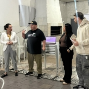 Quatro pessoas estão em pé, lado a lado, em uma sala, durante uma atividade do programa. À esquerda, uma mulher de óculos, blazer branco e calça cinza escuta atentamente. Ao lado dela, um homem de boné preto e camiseta da CUFA RS gesticula com a mão enquanto fala. À direita dele, uma mulher de cabelos pretos compridos observa a conversa, e um homem de moletom bege segura algumas folhas e acompanha a interação. Ao fundo, há cadeiras brancas empilhadas, um computador sobre uma mesa e uma faixa com a identidade visual da CUFA. À direita, uma mesa exibe diversos objetos organizados em fileiras. A cena registra um momento de conversa e troca entre os participantes da atividade.