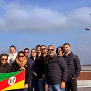 Grupo de visitantes posa ao ar livre segurando a bandeira do Rio Grande do Sul. Ao fundo, aparecem postes de iluminação, céu parcialmente nublado e a paisagem urbana ao longe.