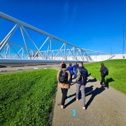 Grupo de visitantes caminha por uma área industrial ao ar livre, ao lado de uma grande estrutura metálica branca. O local tem gramados verdes, céu azul intenso e turbinas eólicas visíveis ao fundo.