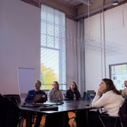Participantes acompanham uma apresentação em sala de reuniões com grandes janelas e iluminação natural. Ao redor de uma mesa, o grupo observa a exposição feita por uma pessoa à frente do ambiente.